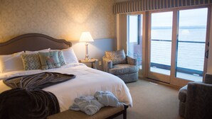 A hotel room with a large bed, a chair, a lamp, and a view of the water through the windows. - Blue Wave Inn (Ashland)