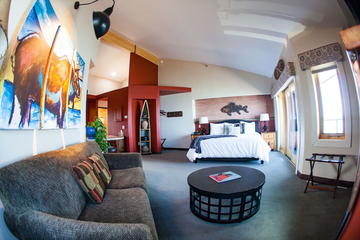 A hotel room with a sofa, a round coffee table, a bed with a headboard, and a large painting on the wall.