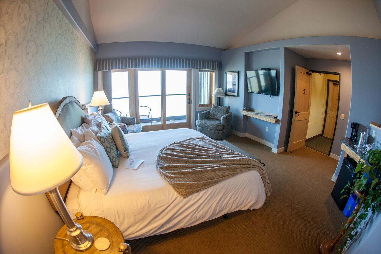 A hotel room with a large bed, a desk with a lamp, a TV, and a sliding door leading to a balcony.