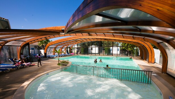 Uma piscina com cobertura de madeira, rodeada por espreguiçadeiras e algumas pessoas.