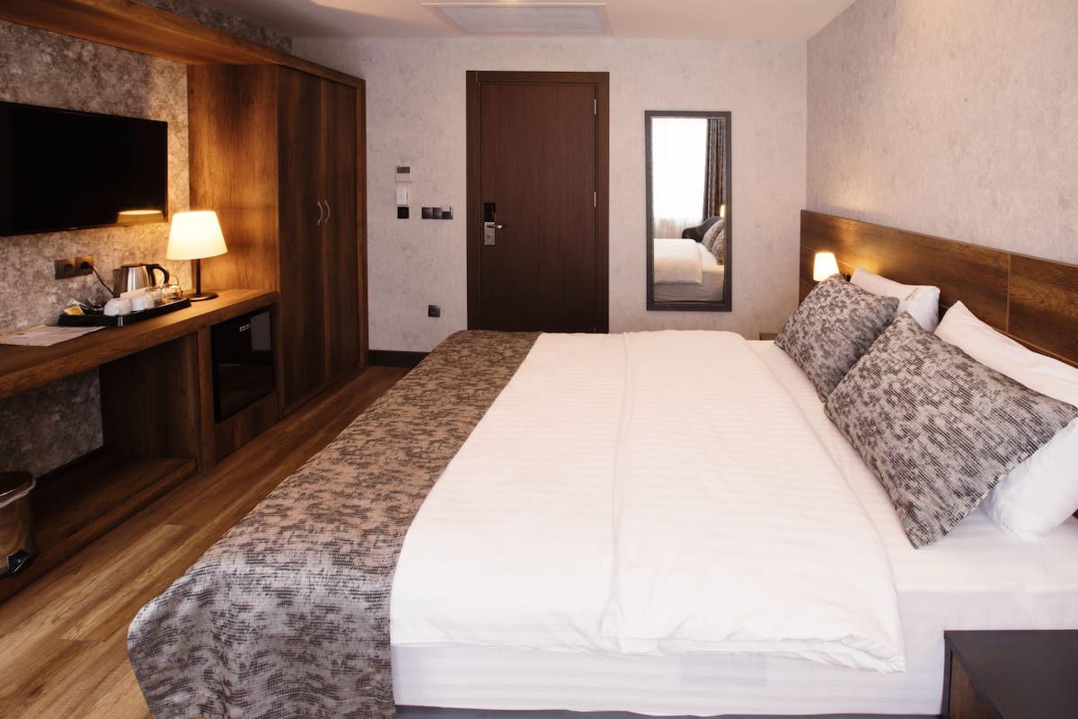 A hotel room with a large bed, a wooden wardrobe, a flat-screen TV, a bedside table with a lamp, and a mirror on the wall.