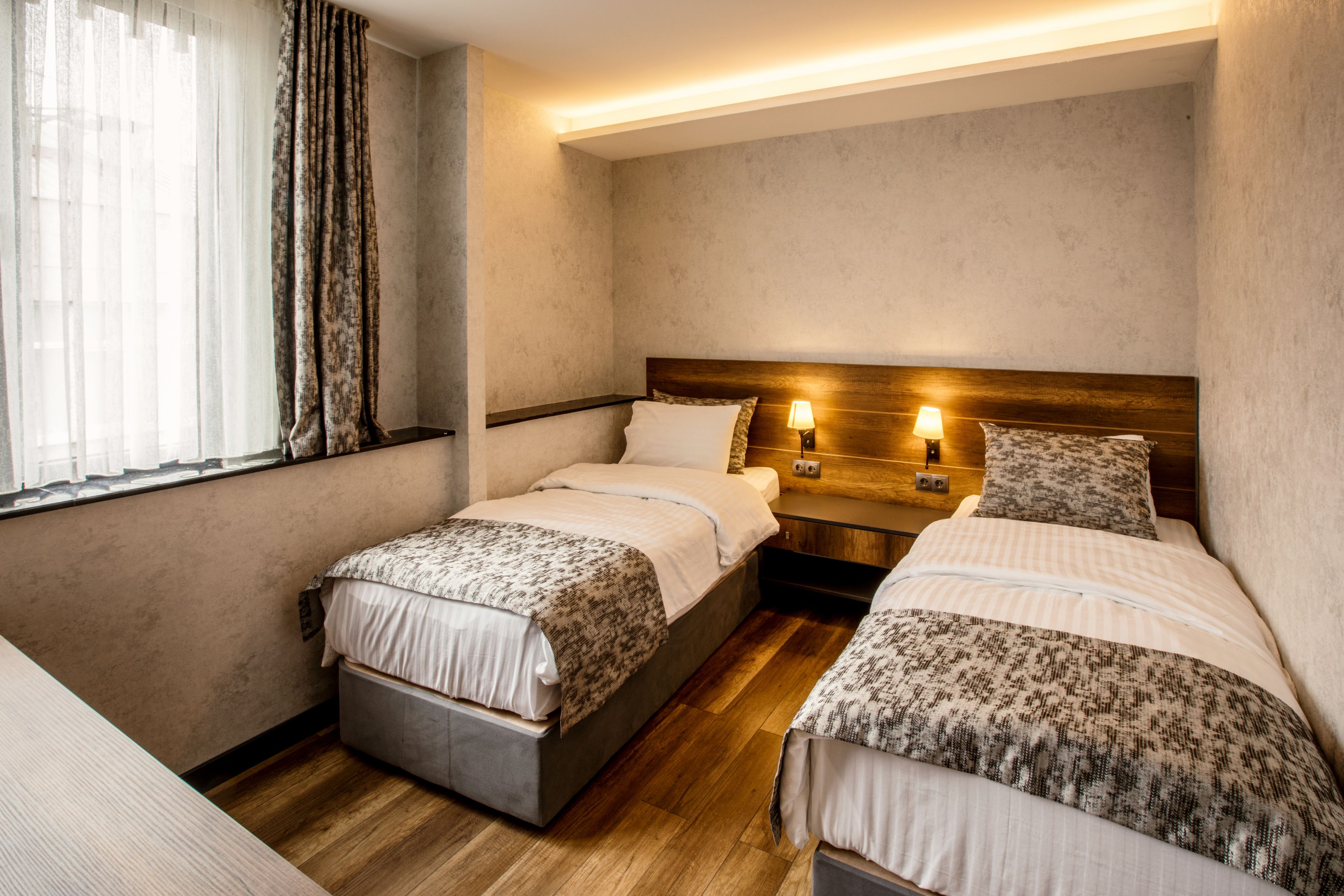 Camera d'albergo con due letti, pavimento in legno, una finestra con tende e un comodino con lampade.