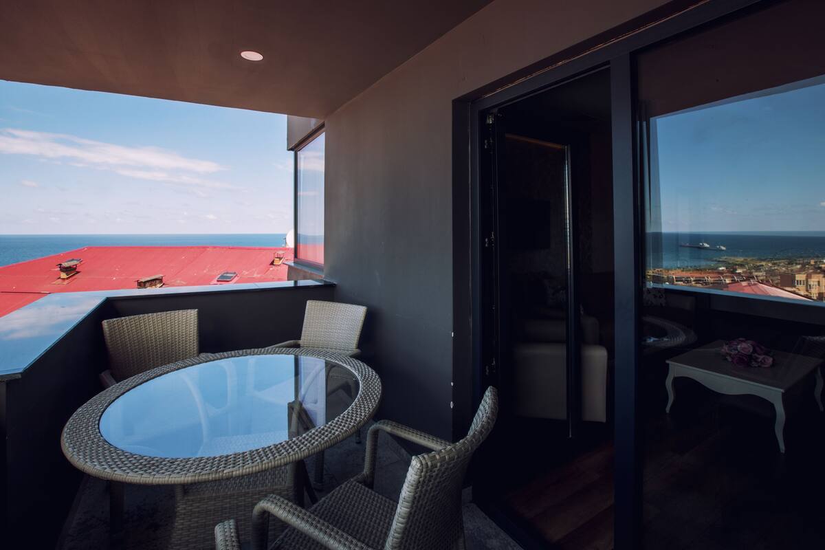 A balcony with a glass table and wicker chairs, overlooking a coastal cityscape.