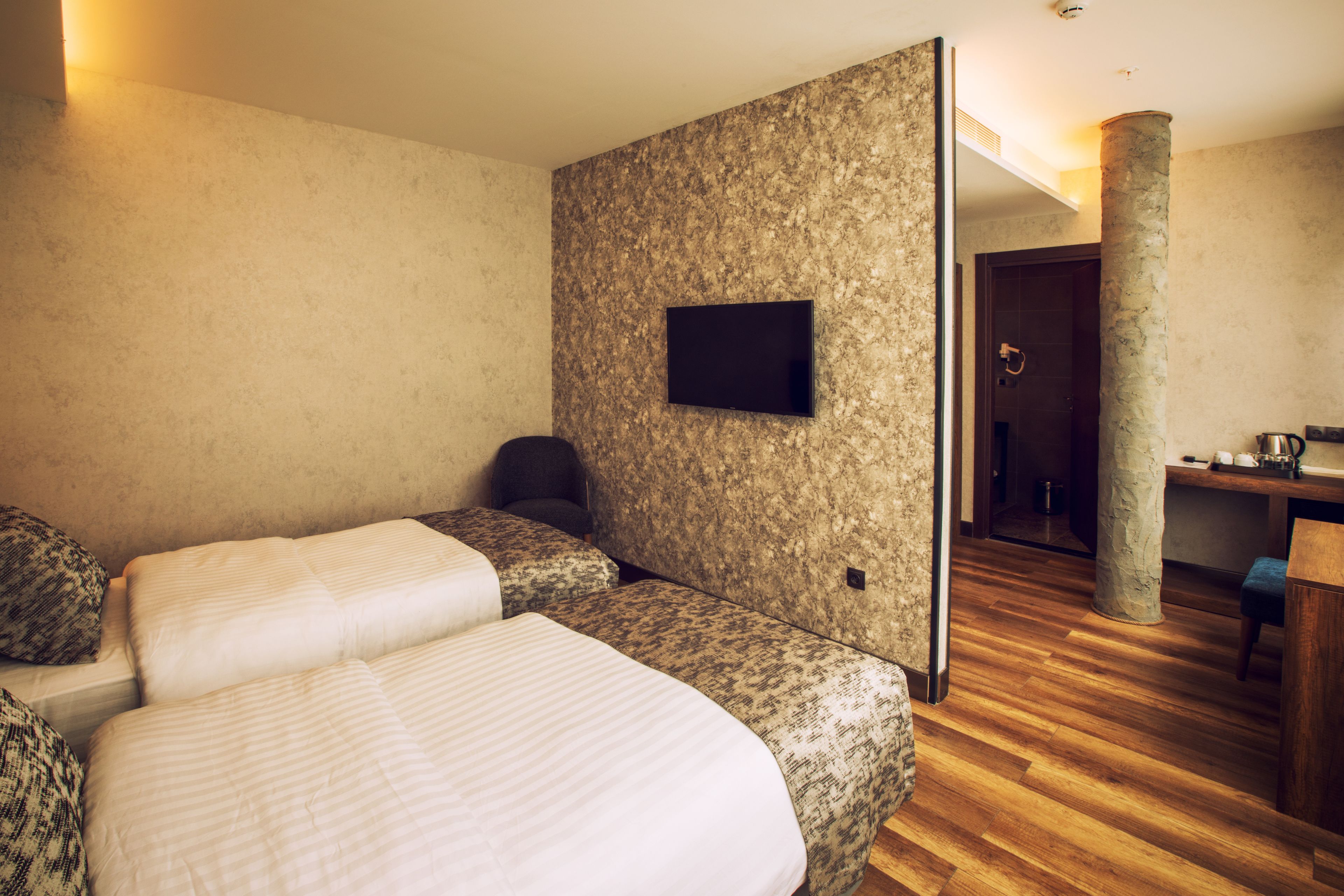 A hotel room with two beds, a television mounted on the wall, a wooden floor, and a bathroom visible through an open door.