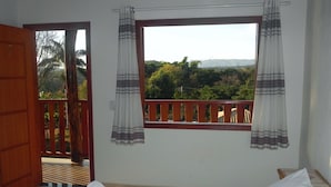 A bedroom with a bed, a window with curtains, a wooden door, and a view of greenery outside. - Pousada Estancia do Vale (Jaboticatubas)