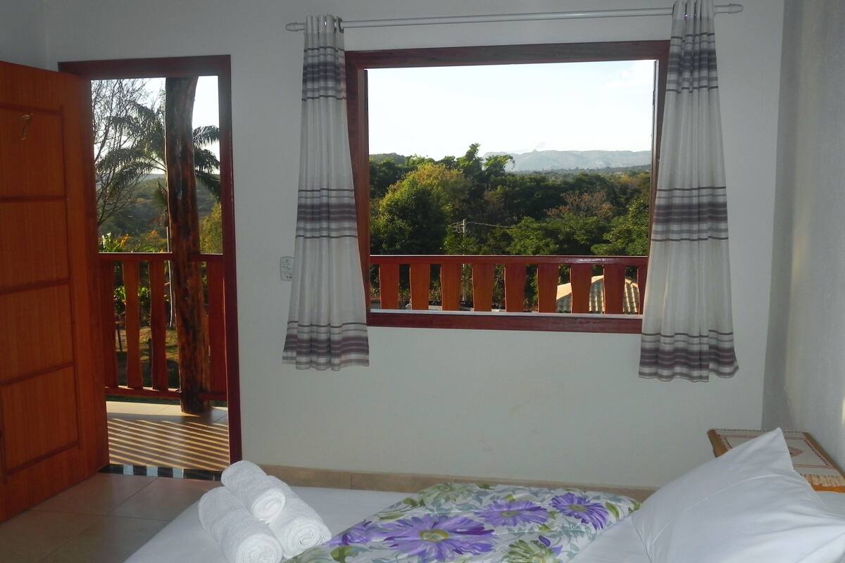 Quarto com cama, janela com cortinas, porta de madeira e vista para o verde lá fora.