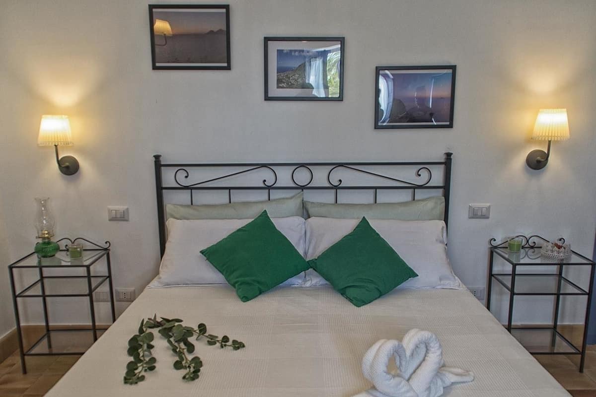 A neatly made bed with green pillows, a white towel, and a green plant on the bed. There are two wall-mounted lamps on either side of the bed.