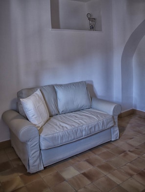 A light grey sofa with a cushion, positioned in a room with a tiled floor and a plain wall.