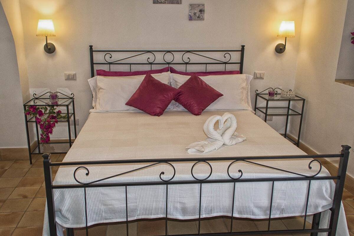 A neatly made bed with a white bedspread, two red pillows, and a white towel arranged in a heart shape. There are two bedside tables with lamps, and a small potted plant on one of the tables.