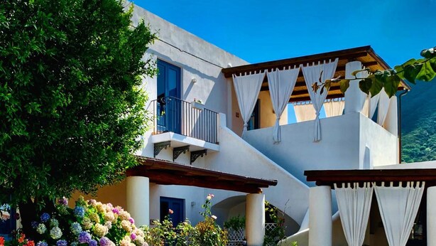 A two-story building with a stone-paved courtyard, white walls, and a balcony with white curtains. - Le sette perle (Malfa)