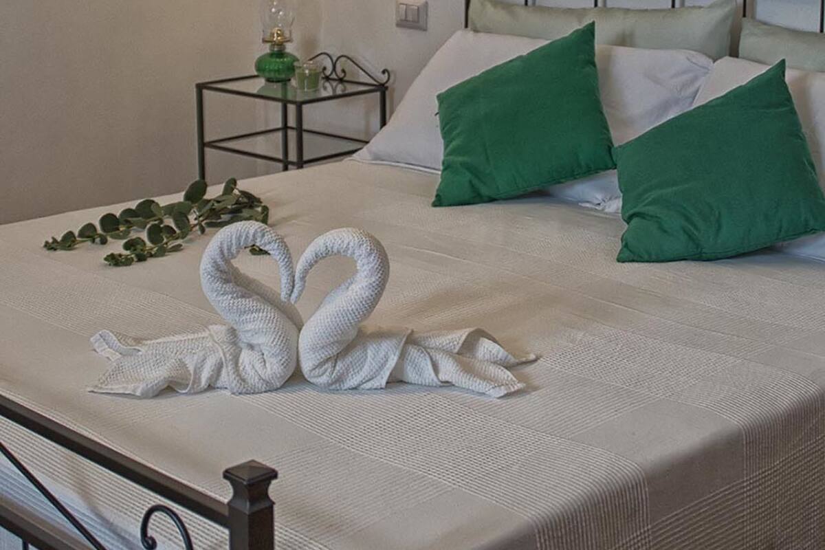 A neatly made bed with a white comforter, two green pillows, and a pair of white towels arranged in a swan shape.