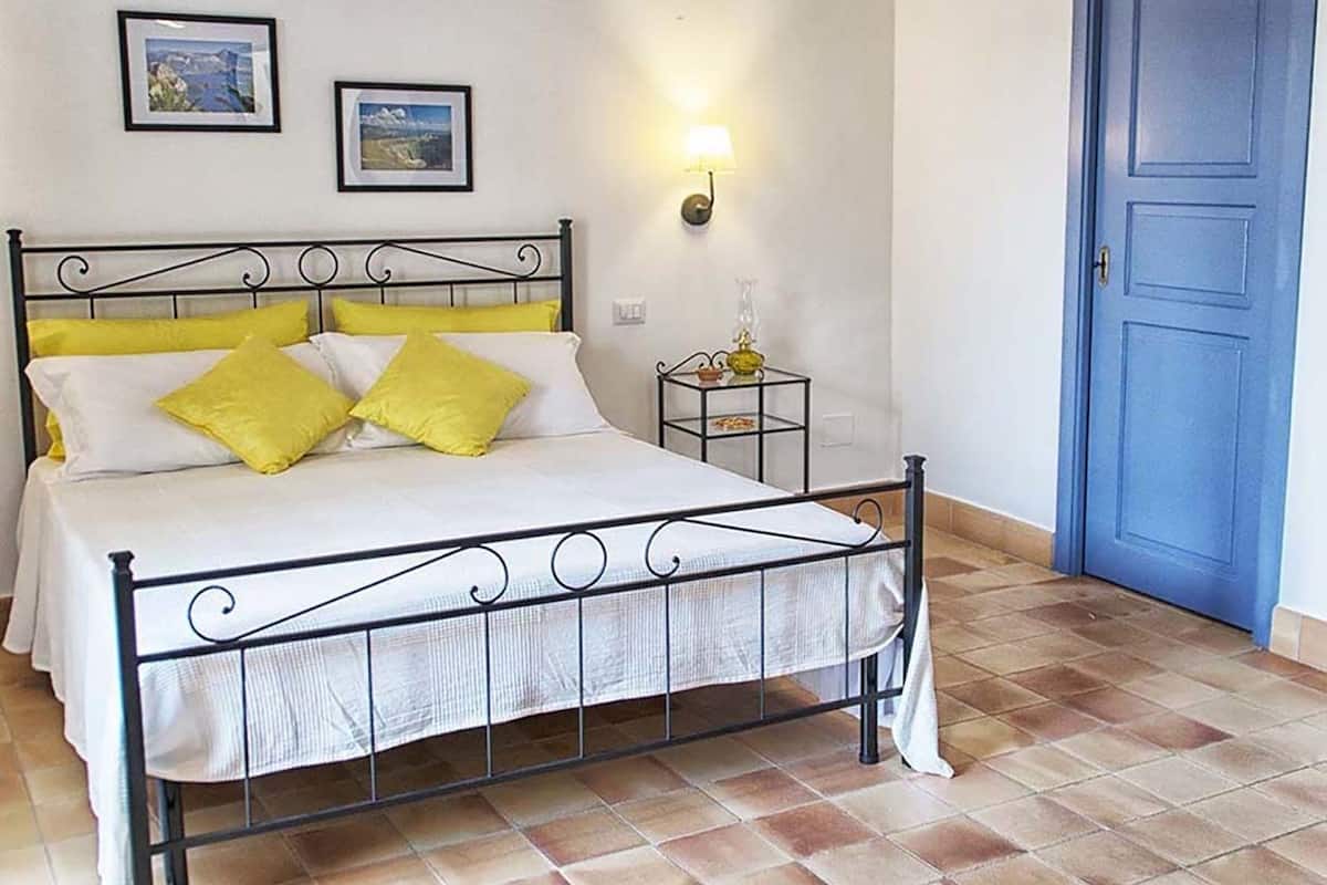 A bed with a metal frame, white bedding, and yellow pillows. There is a small bedside table with a lamp and a picture frame on the wall above the bed.