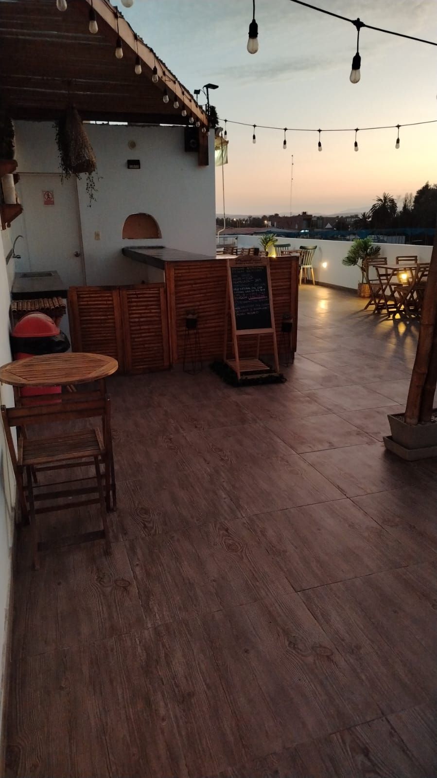An outdoor seating area with wooden tables and chairs, a chalkboard menu, and string lights.