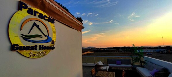 Un letrero de hotel con un logotipo y el texto 'Paracas Guest House Since 2011' sobre un fondo de atardecer.