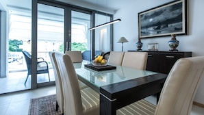 Un comedor con una mesa de vidrio, sillas y un tazón de fruta. Se ve un área exterior con una silla y una mesa a través de puertas de cristal.