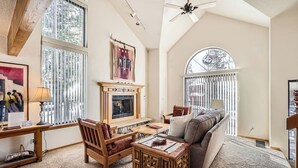 A cozy living room with a fireplace, large windows, and a ceiling fan.
