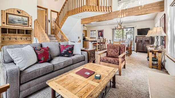 A spacious living area with a grey sofa, patterned cushions, a wooden coffee table, and a red armchair. A wooden staircase with a railing leads to an upper level.