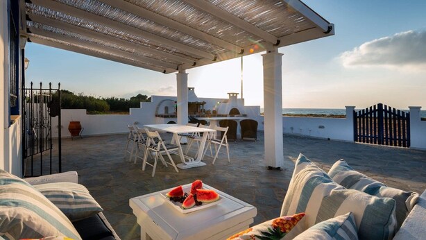 A covered outdoor seating area with a table and chairs, a white table with a watermelon dish, and a view of the sea. - Nautilus Beach House (Rhodes)