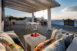 Un área de estar al aire libre cubierta con una mesa y sillas, una mesa blanca con un plato de sandía, y vista al mar.