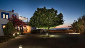 A modern house with a well-lit driveway, a large tree, and a view of the sea at dusk. - Nautilus Beach House (Rhodes)