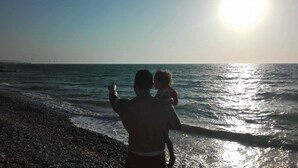 A beach scene with a person holding a child, facing the ocean with waves and a clear sky. - Nautilus Beach House (Rhodes)