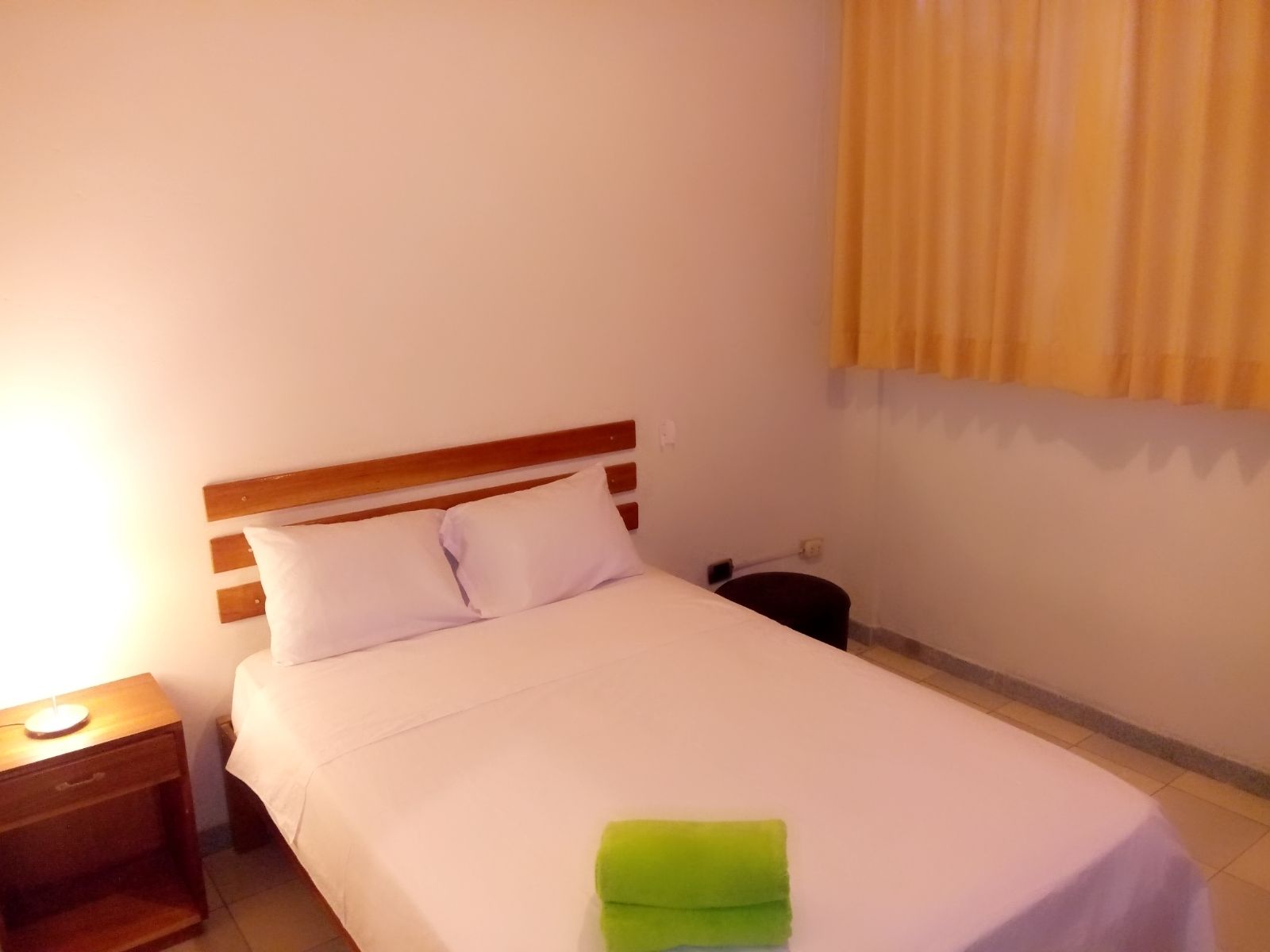 Una cama individual con cabecera de madera, una mesita de noche con lámpara y una toalla verde sobre la cama.