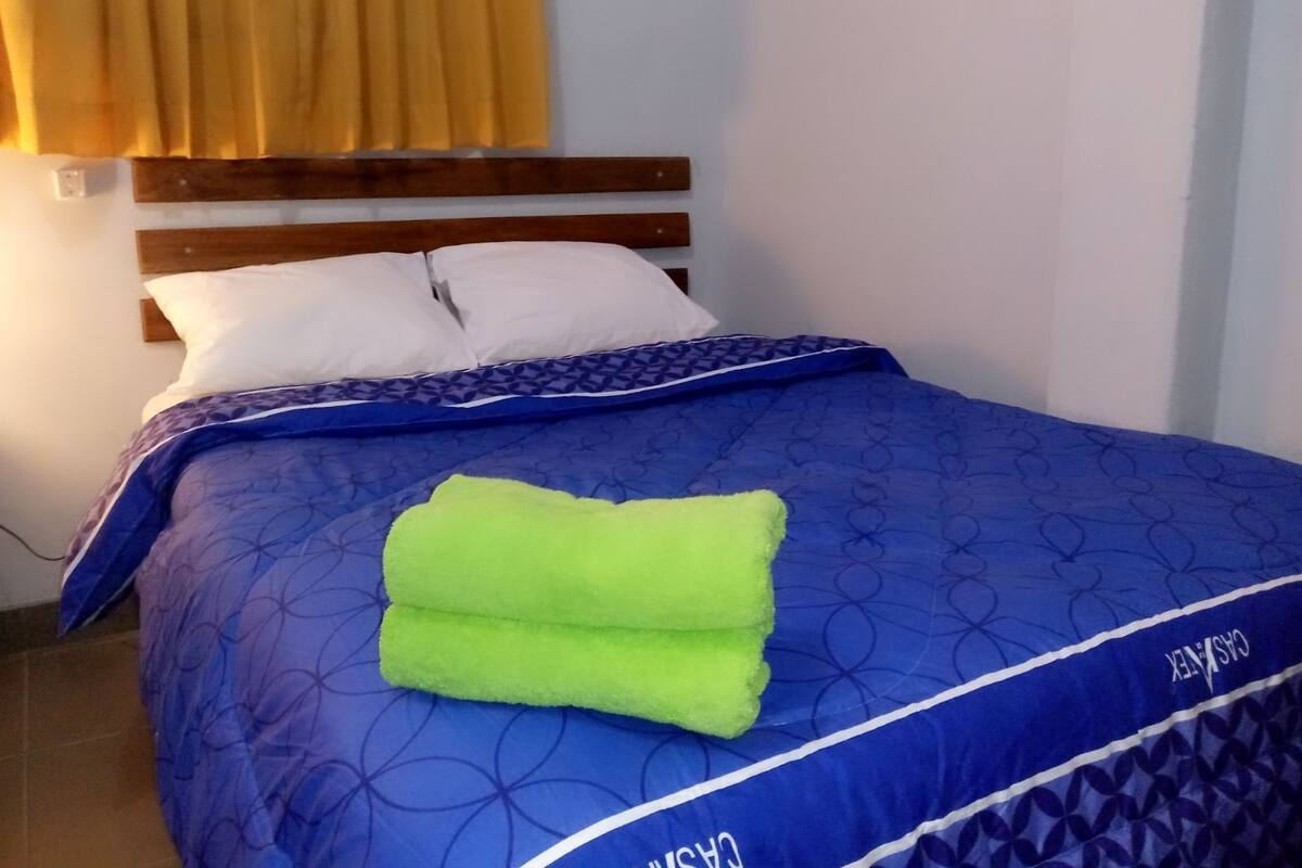 Una cama con colcha azul estampada y almohadas, cabecera de madera y cortina amarilla.