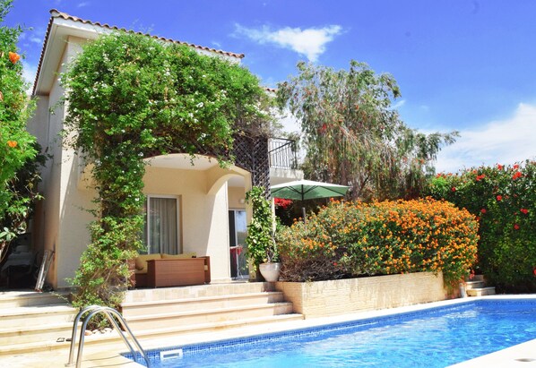 Zona de piscina con escalones que conducen a una casa, rodeada de exuberante vegetación y plantas en flor.
