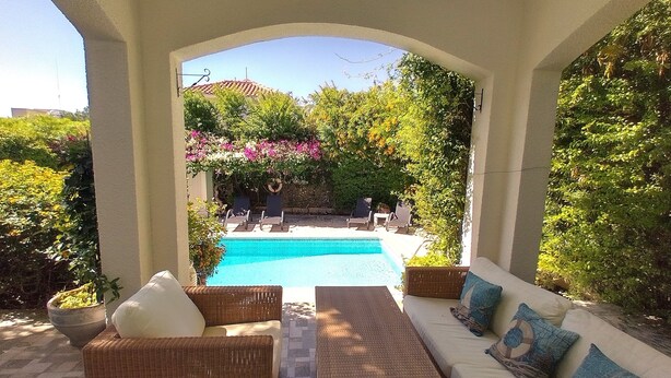 A covered outdoor area with a pool, wicker furniture, and a view of a garden and building in the background. - Villa Talassa near Coral Bay with optional 4x4 CAR, Jacuzzi and HEATED POOL (Paphos)