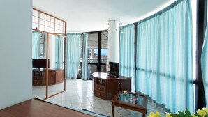 Una sala de estar moderna con una mesa de centro de madera, un televisor sobre un mueble de madera y vistas a través de ventanas grandes con cortinas azules.