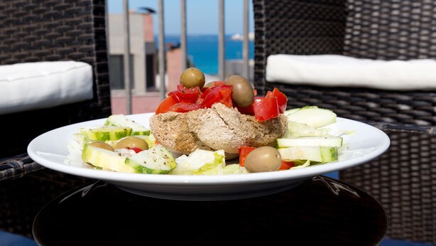 Une assiette de nourriture est posée sur une table, avec une vue sur la mer et une chaise en osier à l’arrière-plan.