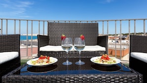 Un balcón con muebles de mimbre, dos copas de vino blanco y platos de comida.