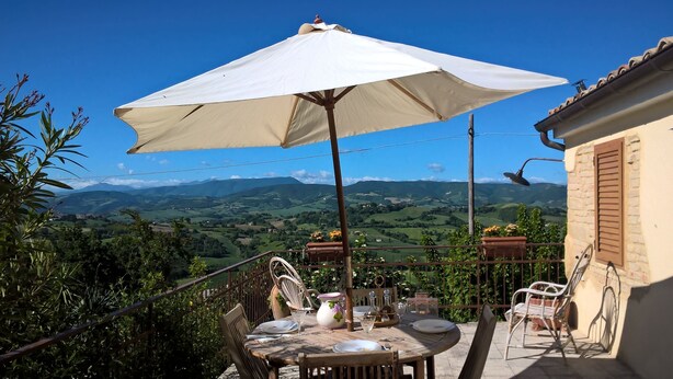 Eine Terrasse mit Esstisch, großem Sonnenschirm und weiter Aussicht auf sanfte Hügel und Berge.