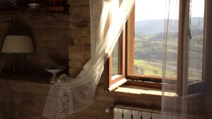 Ein rustikaler Raum mit Steinwand, einem Fenster mit Blick auf ein Tal, einem Heizkörper und einer Lampe auf einem Regal.