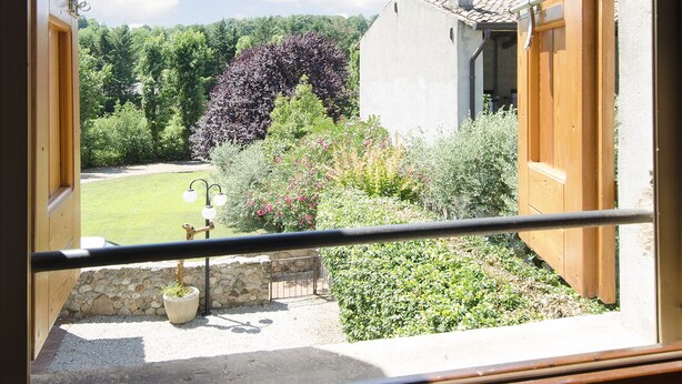 Blick aus einem Fenster mit Balkon auf einen Garten mit verschiedenen Pflanzen und einem kleinen Gebäude.