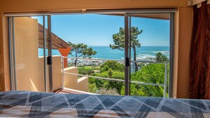Un dormitorio con un amplio ventanal corredera que da a un balcón con vistas al océano y al paisaje urbano.