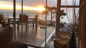 Un balcón con vista al atardecer sobre el océano, terraza de madera y zona de estar al aire libre.