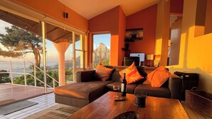 A modern living room with a sofa, a coffee table, and a TV. There is a balcony with a view of the city and a tree. - Exceptional holiday home with panoramic view in Camps Bay (Cape Town)
