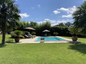 Uma piscina retangular com um deck ladrilhado, rodeada por um gramado bem cuidado e plantas em vasos.