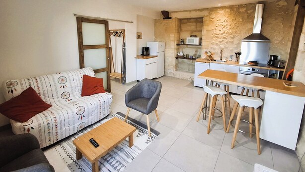 Un espace de vie compact comprenant un canapé, une table basse, un coin repas avec des tabourets, une cuisine équipée d’un four à micro-ondes et un mur en pierre.