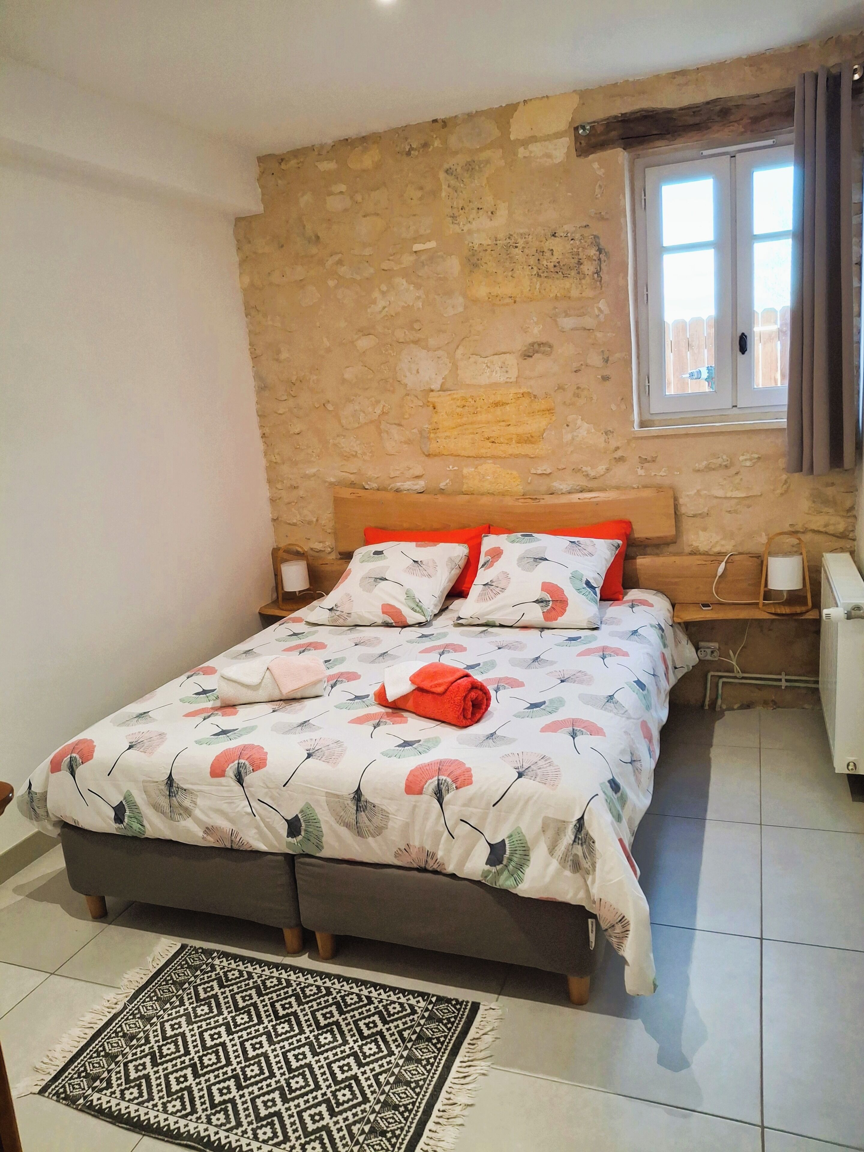 Une chambre avec un mur en pierre, un lit avec une literie à motifs floraux, une tête de lit en bois, une table de chevet, une fenêtre avec des rideaux et un radiateur.