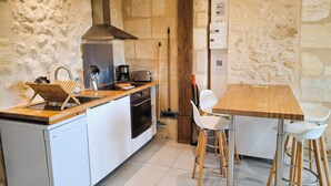 Une cuisine compacte équipée d’un four encastré, d’une machine à café et d’un grille-pain. On y trouve une table en bois avec des chaises blanches, et un balai appuyé contre le mur.