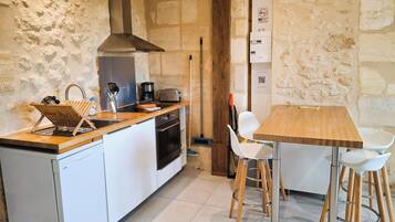 A compact kitchen with a built-in oven, a coffee maker, and a toaster. There is a wooden table with white chairs and a broom leaning against the wall.