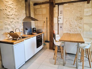 Une cuisine compacte équipée d’un four encastré, d’une machine à café et d’un grille-pain. On y trouve une table en bois avec des chaises blanches, et un balai appuyé contre le mur.