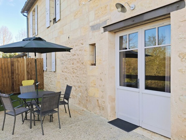 Une terrasse avec une table et des chaises, une clôture en bois et un bâtiment aux murs de pierre et à la porte blanche.