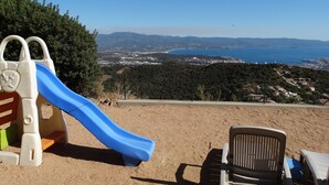 Une aire de jeux avec un toboggan bleu, une structure d’escalade et une vue sur une ville côtière et des montagnes en arrière-plan.