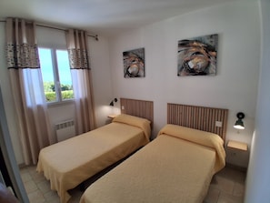 A hotel room with two single beds, a window with curtains, a wall-mounted artwork, and a bedside table with a lamp.