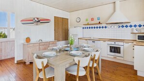 Un coin repas avec une table en bois dressée pour quatre personnes, des chaises blanches et une cuisine dotée de meubles blancs et de carreaux bleus.