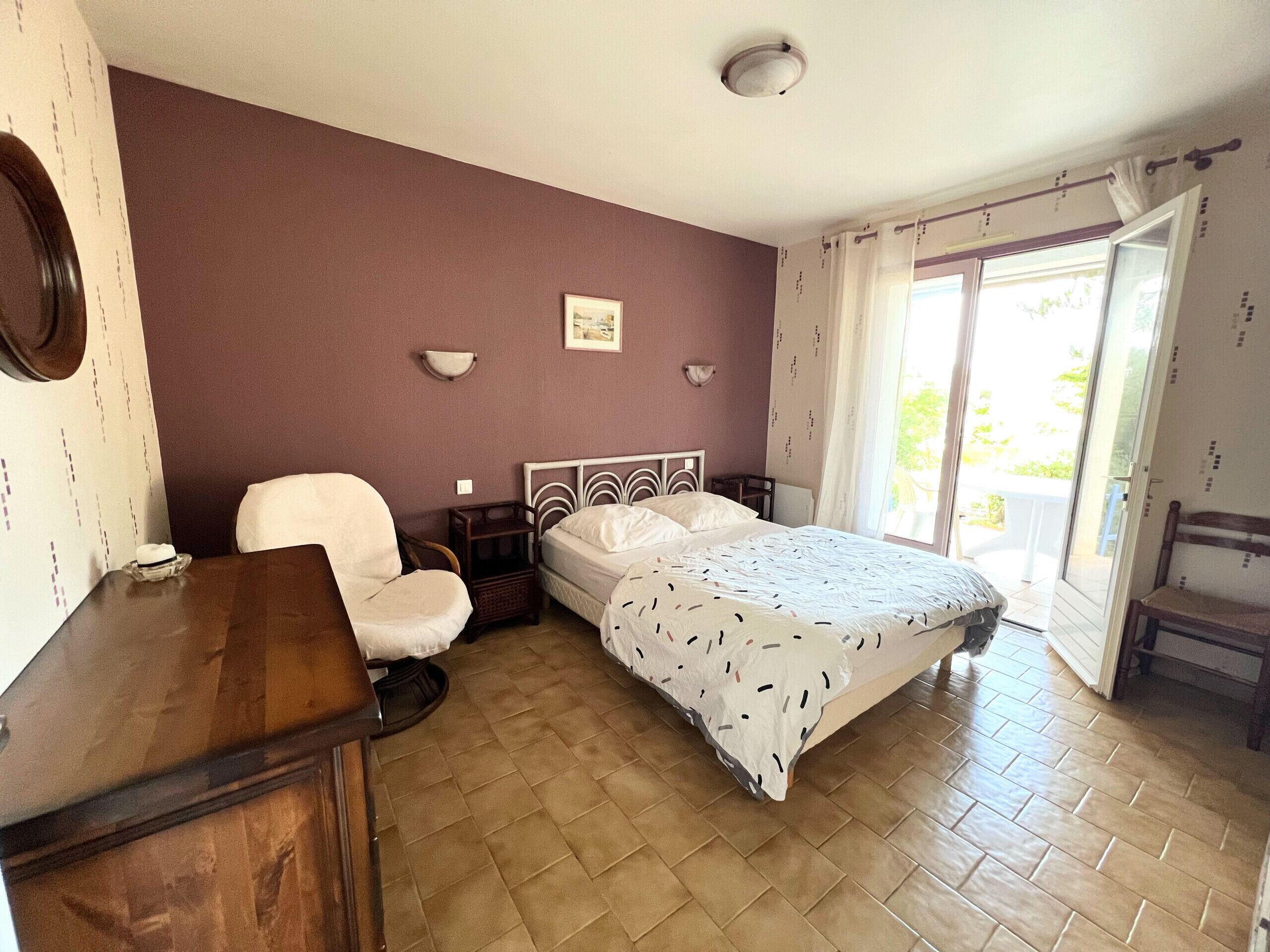 Ein Schlafzimmer mit einem Bett, einem Sessel, einem Nachttisch und einem Fenster mit Vorhängen.