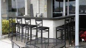 An outdoor bar area with a glass enclosure, multiple seating options including bar stools and a round table, and a view of the cityscape through large windows. - Acropolis Oceanfront Resort (North Wildwood)
